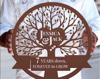 May include: A brown wooden circle with a cutout tree design. The tree has a heart at the center with the names "Jessica & Jack" inside. The banner below reads "7 years down, forever to grow."