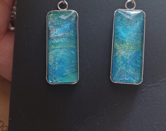 Puede incluir: Un par de pendientes de plata con piezas rectangulares de resina azul y verde. La resina tiene un acabado brillante. Los pendientes están etiquetados con el número "79".