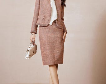 May include: A two-piece women's suit in a muted rose color. The jacket has a round collar, two front pockets, and fringe trim. The matching pencil skirt falls to the knees. A white blouse and cream-colored heels complete the outfit. A small, cream-colored handbag is held in the hand.