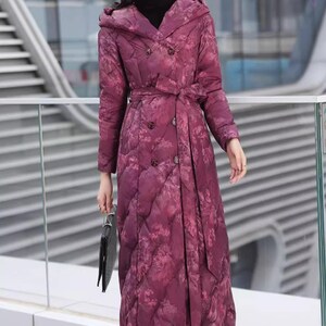 May include: A long, quilted coat in a deep burgundy hue, adorned with a floral pattern. The coat features a hood, double-breasted closure, and a matching belt. The model is wearing black boots and holding a black clutch.