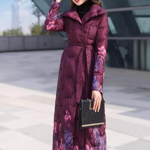 May include: A long, quilted plum-colored coat with a floral pattern in shades of pink and purple. The coat features a belt, a large collar, and long sleeves. A black clutch is held in the hand.