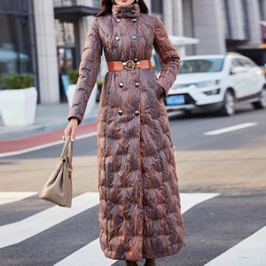 Può includere: Lungo cappotto trapuntato marrone con design doppiopetto e cintura marrone con fibbia dorata. Il cappotto ha un collo alto e viene indossato con stivaletti marroni e una borsa abbinata. Il cappotto ha un motivo unico.