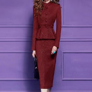 May include: A burgundy two-piece suit with a fitted jacket and a pencil skirt. The jacket features a high collar, button details, and a peplum waist. The skirt is knee-length. The suit has a subtle sparkle and is paired with white heels and a black handbag.