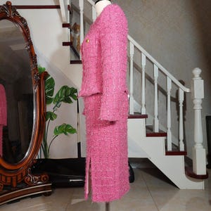 Pu&ograve; includere: Un tailleur gonna in tweed rosa esposto su un manichino. Il completo presenta una giacca con bottoni dorati e una gonna al ginocchio abbinata con uno spacco. Il tessuto ha un aspetto strutturato. Il completo &egrave; esposto davanti a una scala e un grande specchio.