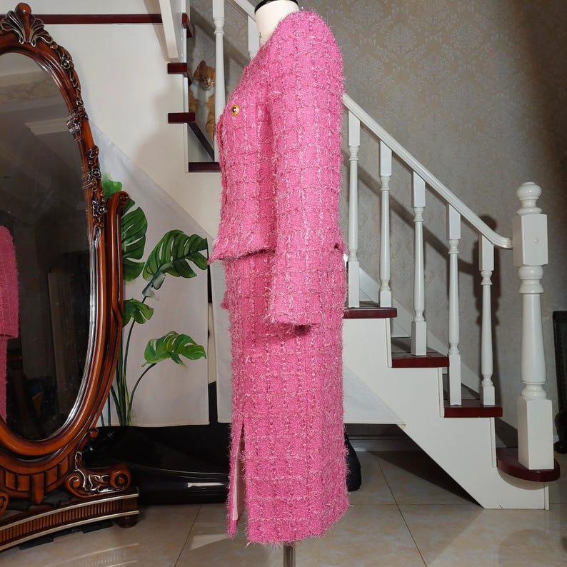 Pu&ograve; includere: Un tailleur gonna in tweed rosa esposto su un manichino. Il completo presenta una giacca con bottoni dorati e una gonna al ginocchio abbinata con uno spacco. Il tessuto ha un aspetto strutturato. Il completo &egrave; esposto davanti a una scala e un grande specchio.