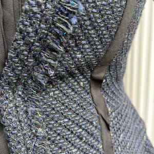 May include: Close-up of a textured, dark blue tweed fabric with frayed edges. The fabric has a woven pattern with flecks of iridescent thread, and is accented by a black trim. The image shows a section of a garment.