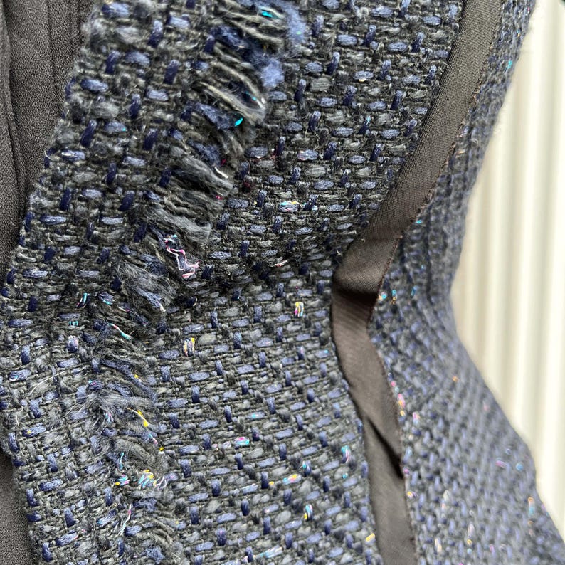 May include: Close-up of a textured, dark blue tweed fabric with frayed edges. The fabric has a woven pattern with flecks of iridescent thread, and is accented by a black trim. The image shows a section of a garment.