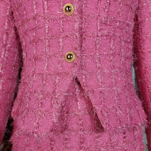 Pu&ograve; includere: Primo piano di un completo in tweed rosa. La giacca ha due bottoni dorati e revers a lancia. La gonna &egrave; visibile sotto la giacca. Il tessuto ha un aspetto strutturato e tessuto con una sottile lucentezza.