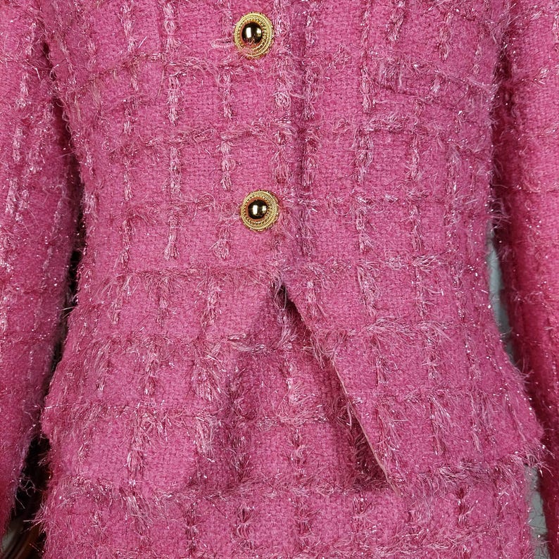 Pu&ograve; includere: Primo piano di un completo in tweed rosa. La giacca ha due bottoni dorati e revers a lancia. La gonna &egrave; visibile sotto la giacca. Il tessuto ha un aspetto strutturato e tessuto con una sottile lucentezza.