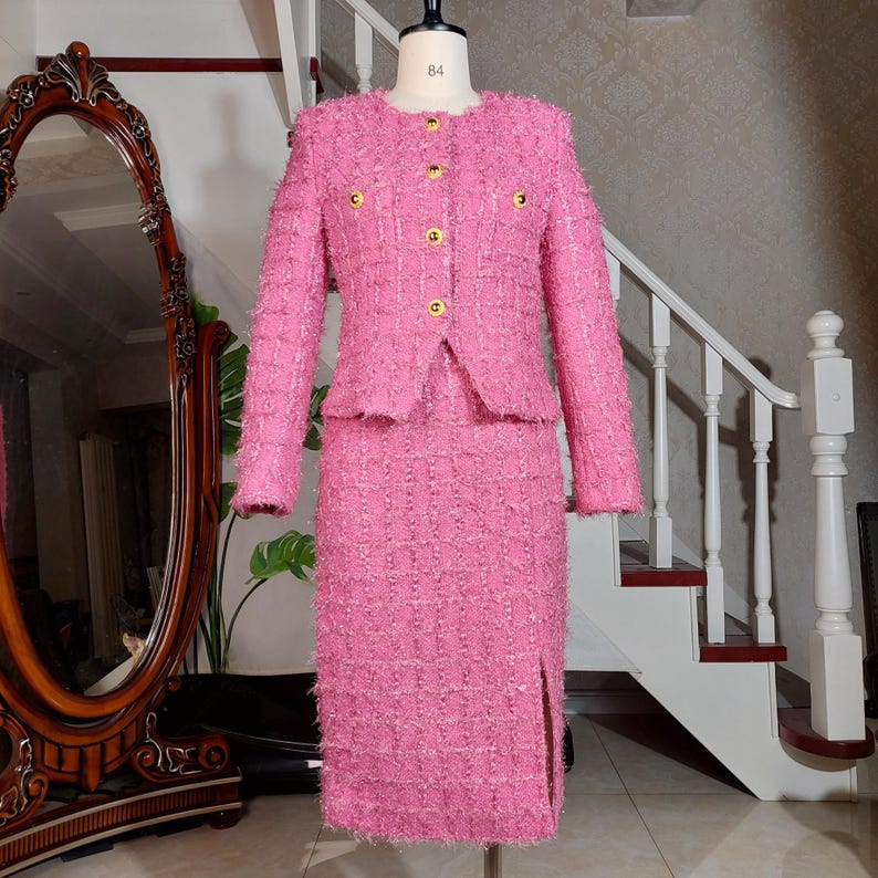 Pu&ograve; includere: Un tailleur gonna rosa in tweed esposto su un manichino. La giacca ha uno scollo tondo, maniche lunghe e bottoni dorati. La gonna &egrave; al ginocchio con uno spacco frontale. Il completo &egrave; realizzato in tessuto strutturato.