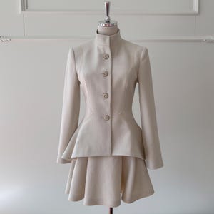 May include: A beige two-piece suit displayed on a mannequin. The suit features a high-collared, button-down jacket with long sleeves and a flared peplum. The matching skirt is a mini-skirt with a flared design. The suit is made of a smooth, neutral-toned fabric.