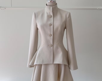 May include: A beige two-piece suit displayed on a mannequin. The suit features a high-collared, button-down jacket with long sleeves and a flared peplum. The matching skirt is a mini-skirt with a flared design. The suit is made of a smooth, neutral-toned fabric.