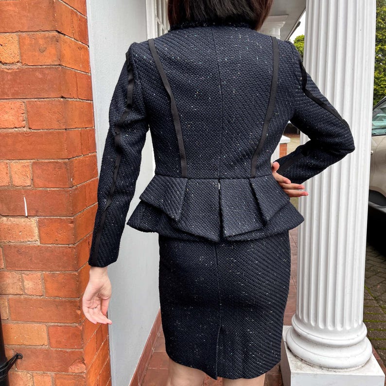 May include: A navy blue tweed skirt suit. The jacket has a peplum waist and black ribbon detailing. The skirt is knee-length. The suit is made of a textured fabric with flecks of white.