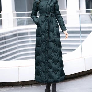 Può includere: Un lungo cappotto imbottito verde scuro con collo alto e cintura abbinata. Il cappotto presenta un design trapuntato e maniche lunghe. Il cappotto è lungo fino al pavimento e ha tasche. La modella indossa stivali neri.