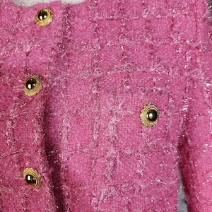 Pu&ograve; includere: Primo piano di una giacca in tweed rosa con bottoni color oro. La giacca ha un aspetto strutturato e tessuto con una sottile lucentezza. I bottoni sono rotondi con un bordo decorativo.