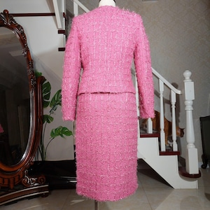 Pu&ograve; includere: Un completo rosa in tweed a due pezzi, composto da una giacca a maniche lunghe e una gonna al ginocchio. Il completo &egrave; esposto su un manichino, mettendo in risalto il tessuto strutturato e il design su misura. Il completo &egrave; di una tonalit&agrave; rosa vibrante.