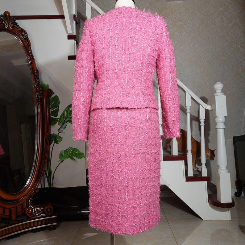 Pu&ograve; includere: Un completo rosa in tweed a due pezzi, composto da una giacca a maniche lunghe e una gonna al ginocchio. Il completo &egrave; esposto su un manichino, mettendo in risalto il tessuto strutturato e il design su misura. Il completo &egrave; di una tonalit&agrave; rosa vibrante.