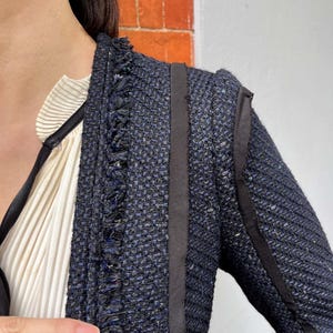 May include: Close-up of a navy blue tweed jacket with black trim and fringe detailing. The jacket has a textured weave and a tailored fit. A cream-colored blouse with a pleated design is visible underneath the jacket.