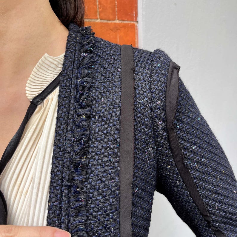 May include: Close-up of a navy blue tweed jacket with black trim and fringe detailing. The jacket has a textured weave and a tailored fit. A cream-colored blouse with a pleated design is visible underneath the jacket.