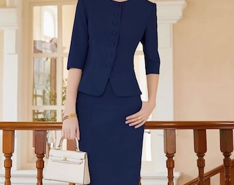 Puede incluir: Un traje de falda azul marino con una chaqueta entallada con cuello redondo y cierre de botones, combinada con una falda lápiz a la rodilla a juego. La chaqueta tiene mangas tres cuartos. Se sostiene un bolso blanco en la mano.
