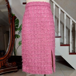 Pu&ograve; includere: Gonna a tubino in tweed rosa con spacco frontale. La gonna &egrave; esposta su un manichino. Il tessuto ha un aspetto strutturato e tessuto. La gonna &egrave; lunga fino al ginocchio e ha un design classico e su misura.