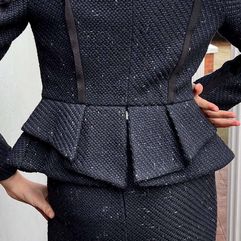 May include: A dark navy blue tweed suit with a peplum jacket and a matching skirt. The jacket features a fitted silhouette with a layered peplum detail and black ribbon trim. The fabric has a textured appearance with flecks of white.