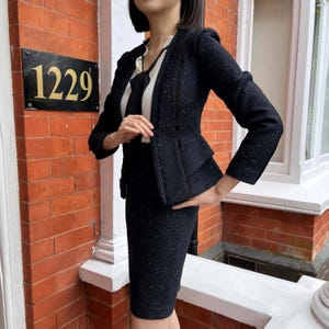 May include: A dark navy blue skirt suit with a textured weave. The jacket has a tailored fit with a peplum detail and a contrasting trim. The skirt is knee-length. The outfit is worn over a white blouse with a black tie.