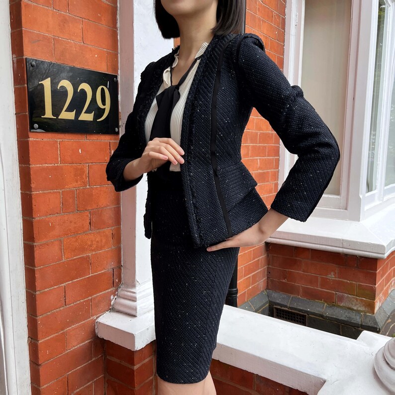 May include: A dark navy blue skirt suit with a textured weave. The jacket has a tailored fit with a peplum detail and a contrasting trim. The skirt is knee-length. The outfit is worn over a white blouse with a black tie.