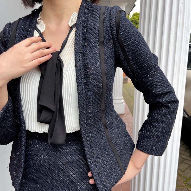 May include: A navy blue tweed blazer with black trim and a matching skirt. The blazer has a textured weave and a fringe detail along the edges. A white blouse with a black bow is worn underneath. The outfit is a classic, elegant style.
