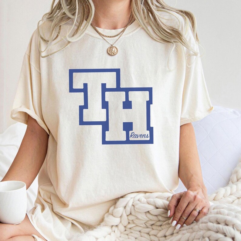 May include: Cream-colored t-shirt with a blue graphic featuring the letters "TH" inside a blue rectangle. Below the letters, the word "Ravens" is printed in blue. The shirt has short sleeves and a relaxed fit.