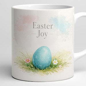 Könnte beinhalten: Weiße Keramik-Tasse mit Ostermotiv. Die Tasse zeigt ein blau gesprenkeltes Ei, grünes Gras und kleine rosa und weiße Blumen. Die Worte "Easter Joy" sind in einer weichen grauen Schrift auf die Tasse gedruckt.