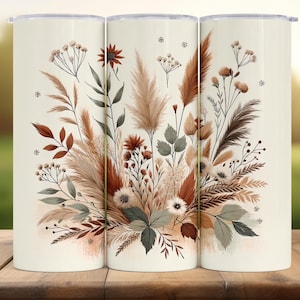 Op de afbeelding: Set van drie witte geïsoleerde bekers met een bloemenontwerp. Het ontwerp toont een aquarel illustratie van diverse gedroogde bloemen en bladeren in bruin, beige en groen. De bekers zijn cilindrisch en hebben een zilveren deksel.