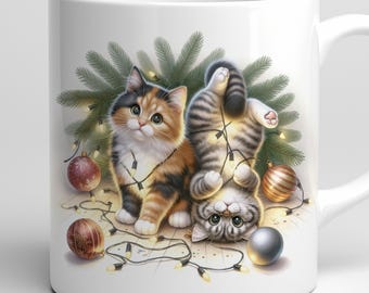 May include: White ceramic mug featuring two playful kittens amidst Christmas decorations. One kitten is orange, black, and white, while the other is gray and white. Both are tangled in string lights, with ornaments and a fir branch in the background.