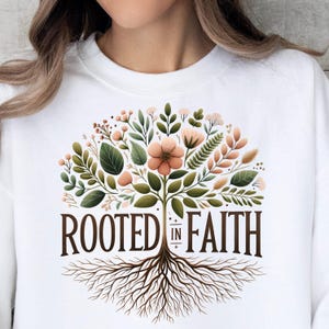 A white t-shirt with a graphic design featuring a tree with roots and flowers, along with the text "ROOTED IN FAITH".