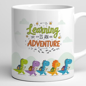 May include: White ceramic mug featuring the phrase "Learning is an Adventure" in green and orange. The mug showcases a line of colorful cartoon dinosaurs, each carrying a backpack and a notepad, with a trail of footprints below.