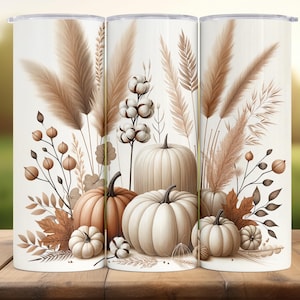 Op de afbeelding: Set van drie witte geïsoleerde bekers met een herfstthema. Het ontwerp bevat pompoenen, katoen en tarwearen in bruine en beige tinten, wat een rustieke uitstraling creëert. De bekers zijn cilindrisch en hebben een glanzende afwerking.