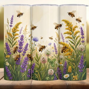 Puede incluir: Tres vasos de acero inoxidable con un diseño envolvente que presenta abejas, lavanda y flores silvestres. El diseño incluye elementos amarillos, morados, verdes y blancos. Los vasos son cilíndricos y tienen una tapa plateada.