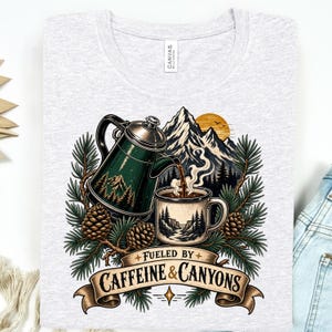 May include: A light grey heather t-shirt featuring a graphic of a green coffee percolator pouring coffee into a mug, surrounded by pine branches, pinecones, and mountains. Below the illustration is a banner with the text FUELED BY CAFFEINE & CANYONS.