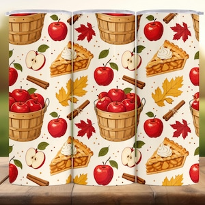 May include: A cylindrical tumbler featuring a repeating pattern of red apples, wooden bushel baskets filled with apples, slices of apple pie topped with whipped cream, cinnamon sticks, and autumn leaves in shades of yellow and red on a cream background.