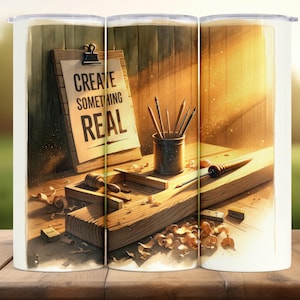 May include: A white tumbler featuring a watercolor illustration of a woodworking scene. The image includes a clipboard with the text "CREATE SOMETHING REAL", pencils, and woodworking tools on a wooden surface. The background is a warm, sunlit workshop.