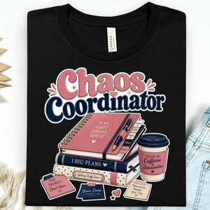 May include: A black t-shirt featuring the text Chaos Coordinator in pink and navy blue. The graphic includes a stack of three notebooks, a pen, a coffee cup with the text Fueled by Caffeine & Determination, and several sticky notes with text.