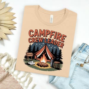 Puede incluir: Una camiseta color beige con la frase "Campfire Crew Leader" en naranja y blanco. El diseño incluye una fogata, una tienda de campaña, árboles y un letrero de "Basecamp". La camiseta está doblada sobre una superficie blanca.