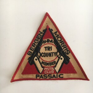 May include: A triangular patch with a red and tan border. The patch features the text "Bergen Morris" and "Passaic" with the words "Tri County Member Pistol Club" in the center. Two black pistols are on the patch.