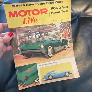 May include: A vintage "Motor Life" magazine cover from February 1956, featuring a green Ford V-8 car. The cover has the text "What's New in the 1956 Cars" and "FORD V-8 Road Test". The magazine is held by a person.