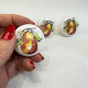May include: Three round white ceramic cabinet knobs, each featuring a detailed watercolor-style illustration of a yellow and brown pear with green leaves and small white blossoms. The knobs have a smooth, glossy finish and a circular shape.