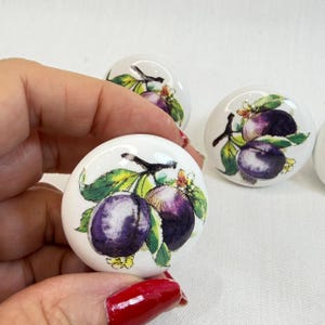May include: A set of round white ceramic cabinet knobs featuring a detailed watercolor-style illustration of dark purple plums with green leaves and small yellow flowers. The design is printed on the surface of each smooth, circular knob.