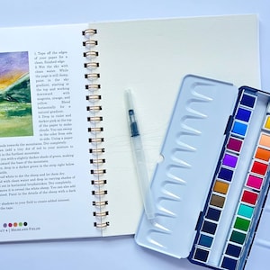 May include: An open spiral-bound sketchbook with a watercolor painting of a mountain landscape. A watercolor paint set with 24 colors and a water brush are next to the sketchbook. The painting is of a mountain with green and purple hues.