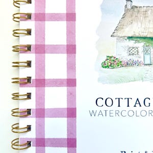 May include: A pink and white gingham notebook with a gold spiral binding. The cover features a watercolor illustration of a cottage with a thatched roof and a blue door. The text "Cottage Watercolor" and "Paint & Studio" is printed on the cover.