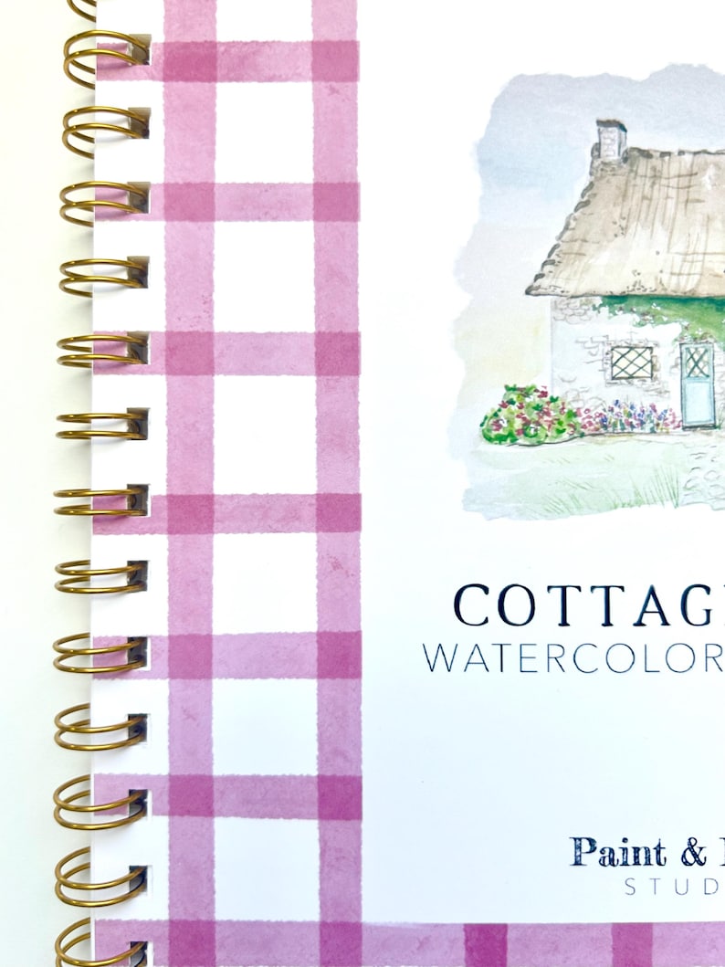 May include: A pink and white gingham notebook with a gold spiral binding. The cover features a watercolor illustration of a cottage with a thatched roof and a blue door. The text "Cottage Watercolor" and "Paint & Studio" is printed on the cover.