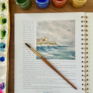 Puede incluir: Un diario de arte abierto con una acuarela de una escena costera, un pincel y peque&ntilde;os botes de pintura. El texto de la p&aacute;gina dice "Project 9 | THE COUNT OF MONTE CRISTO".